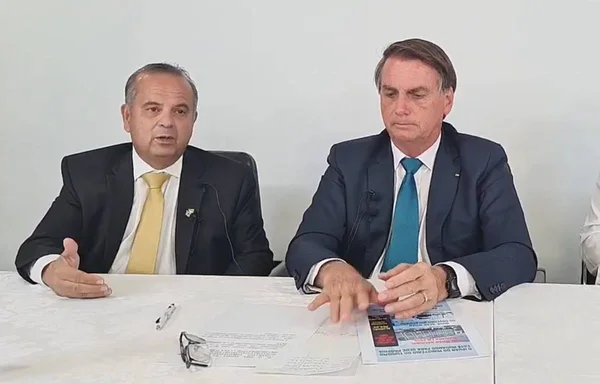 O ministro do Desenvolvimento Regional, Rogério Marinho, aparece ao lado do presidente Bolsonaro em meio a fala. Ambos possuem microfones a frente, sentados numa mesa branca, com folhas de papel espalhadas - Metrópoles