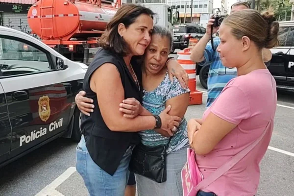 Foto colorida mostra uma delegada, vestindo colete preto, abraçando uma mulher negra vestindo camisa azul clara estampada. Ao lado está mulher com camiseta rosa e coque no cabelo.