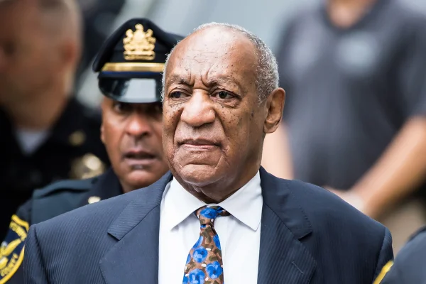 Foto colorida de Bill Cosby usando terno e acompanhado de um policial