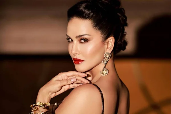 Foto colorida da atriz pornô Sunny Leone de costas olhando para a câmera, de batom vermelho e cabelo preso - Metrópoles