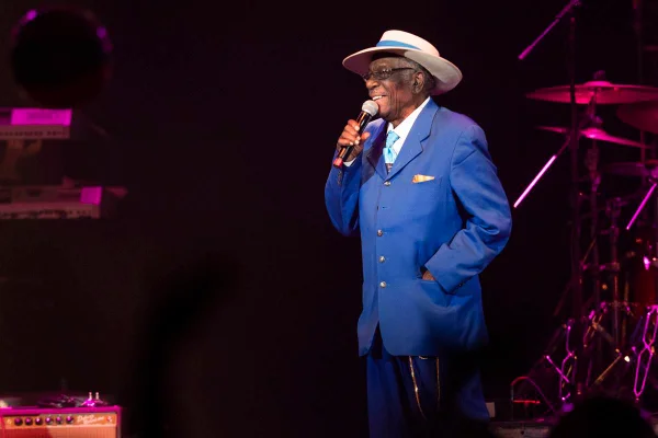 foto colorida de homem idoso negro de terno azul e chapeu cantando - metrópoles