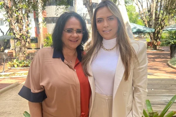 Fotografia colorida de duas mulheres com roupas claras sorrindo para foto lado a lado