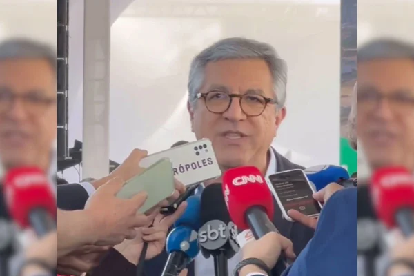 Homem branco de cabelos brancos usa óculos de aro redondo, ele responde a jornalistas que colocam microfones e celulares na frente dele - Metrópoles