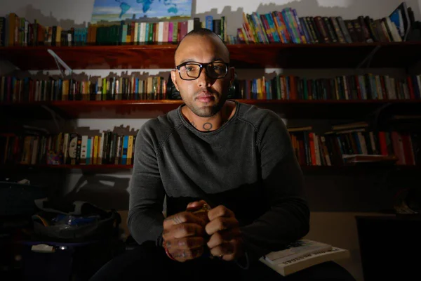 Foto colorida de homem negro em quarto com livros e mãos machucadas