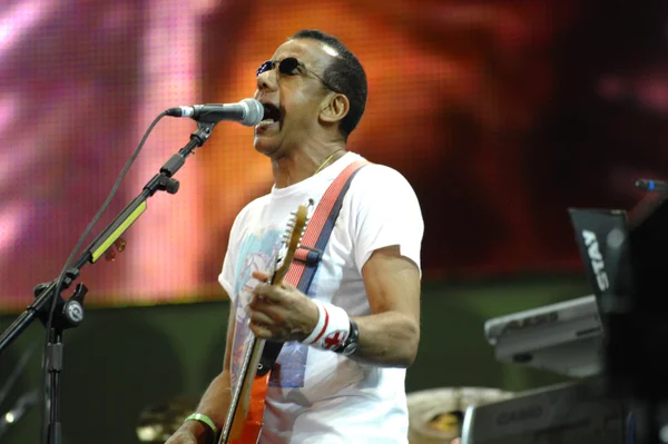 imagem colorida mostra cantor jorge ben jor, ele é negro, tem o cabelo curto, veste óculos escuros e uma blusa branca. jorge segura um instrumento de cordas e canta em frente em um microfone - metrópoles