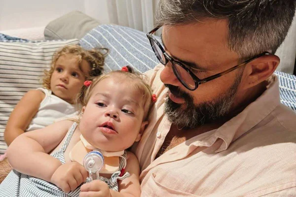 foto colorida de Juliano Cazarré com Maria Guilhermina no colo e outra filha deitada ao lado - metrópoles