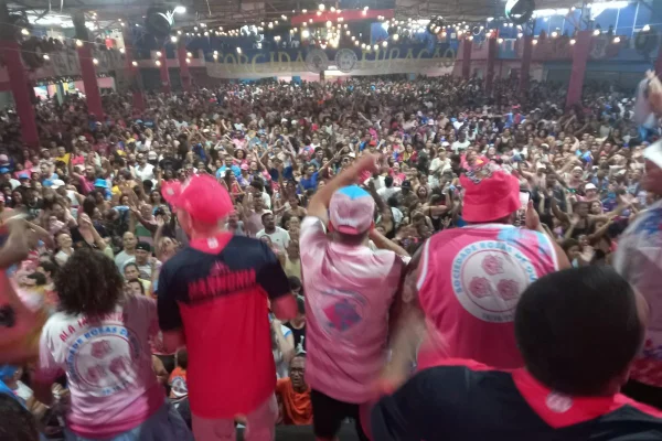 Imagens coloridas mostram grupo de pessoas sobre um mezanino, com camisetas rosas, cores da escola de samba Rosas de Ouro, e multidão ao fundo de uma quadra de esportes, em comemoração ao título do carnaval paulistano de 2025