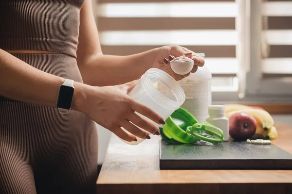 Mulher atlética em roupas esportivas com colher de medição na mão coloca porção de proteína de soro de leite em pó em um shaker em mesa de madeira com cápsulas brancas de aminoácidos, bananas e maçã, fazendo bebida proteica - Metrópoles