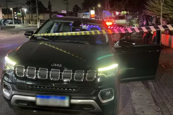 Imagens coloridas mostram carro da marca Jeep parado após assalto, faróis acessos, atrás de uma faixa de isolamento usada pela polícia