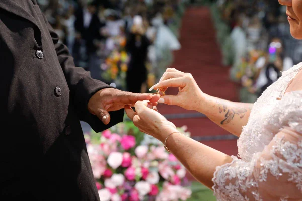 Foto colorida de casal, de casal, homem e mulher, de terno e vestido de noiva, em altar colocando a aliança no dedo do outro