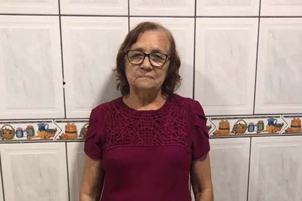 Mulher idosa com blusa vinho e óculos em frente a parede