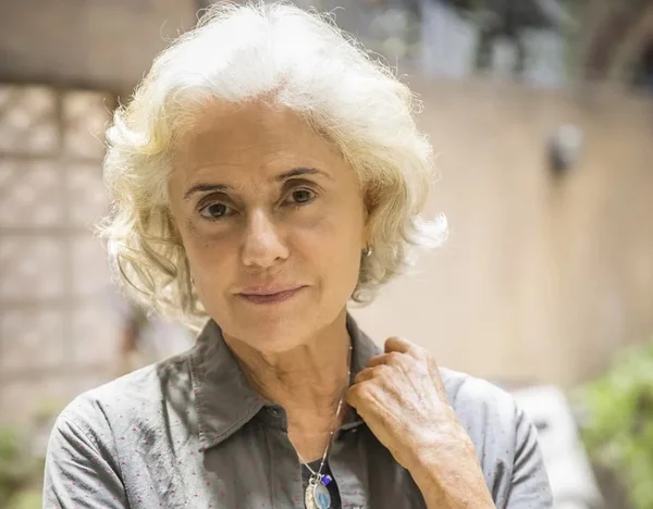 Na imagem colorida, a atriz Marieta Severo está centralizada. Ela veste blusa cinza, tem cabelos brancos e está com a mão esquerda sobre o ombro esquerdo. Ela sorri timidamente para a câmera