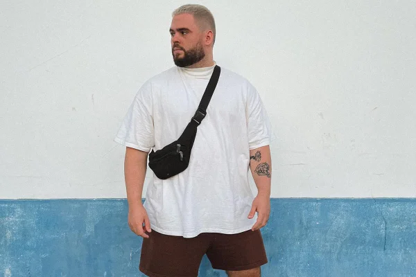 Homem branco e jovem, de barba preta e cabelo loiro descolorido curto, posa para foto em frente a um muro na rua. Ele usa uma camiseta branca, uma pochete preta cruzada no colo e uma bermuda marrom. - Metrópoles