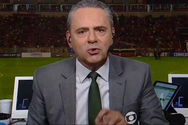 Foto de um homem branco em uma cabine de narração. Ele tem cabelo preto com grisalhos, usa um terno e ao fundo um campo de futebol - Metrópoles
