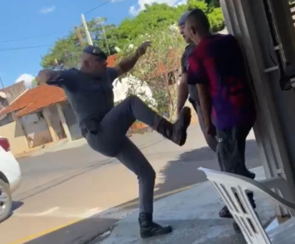 Imagem colorida mostra policial militar dando chute em suspeito de roubo, ue aparece com camiseta roxa e rosa