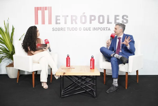 Fotografia colorida mostrando repórter entrevistando homem de terno, ambos sentados-Metrópoles