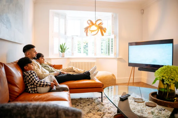 Foto mostra Pai e dois filhos relaxando em um sofá de couro marrom em uma sala de estar moderna, assistindo à TV e aproveitando um fim de semana tranquilo em família, em um lar confortável e multiétnico. - TV 3.0