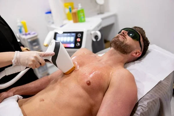 Home branco e sem camisa deitado em uma maca enquanto uma outra pessoa passa uma máquina parecida com uma pistola em seu peito