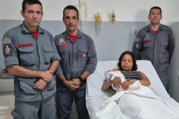 Imagem colorida com bombeiros ao lado de uma mulher segurando um recém-nascido. A mãe está deitada enquanto os policiais estão em pé.