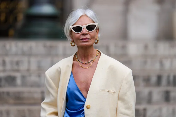Mulher idosa branca, com cabelos curtos e também brancos, com roupa estilosa. Ela usa um óculos de sol vintage bege, um colar de pérolas multicoloridas, um colar de corrente de ouro, um blazer largo branco, um colete de seda azul brilhante com decote em V abotoado.