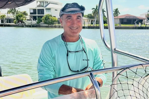Foto colorida de homem sorrindo para foto, ele está em um barco enquanto veleja em uma espécie de rio/lago - Metrópoles.