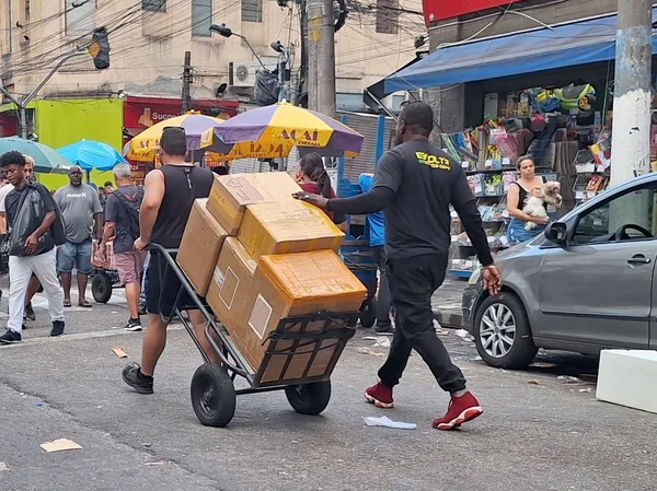 Imagem mostra homem puxando carrinho de mão, seguido por outro, em meio a uma rua com outras pessoas - Metrópoles