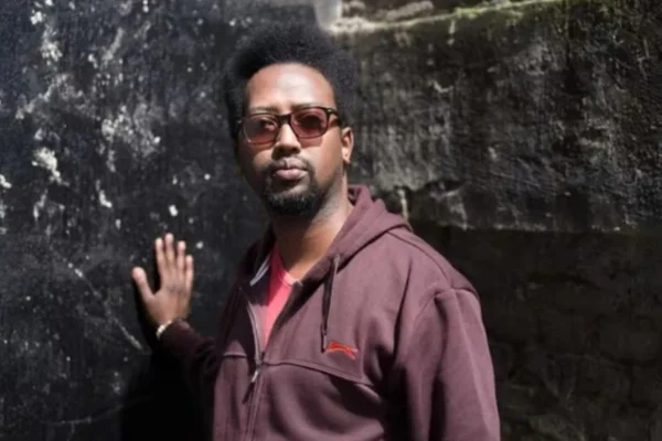 Imagem mostra Alpha Kabeja, que é um homem negro e está com casaco roxo, encostado em uma parede - Metrópoles