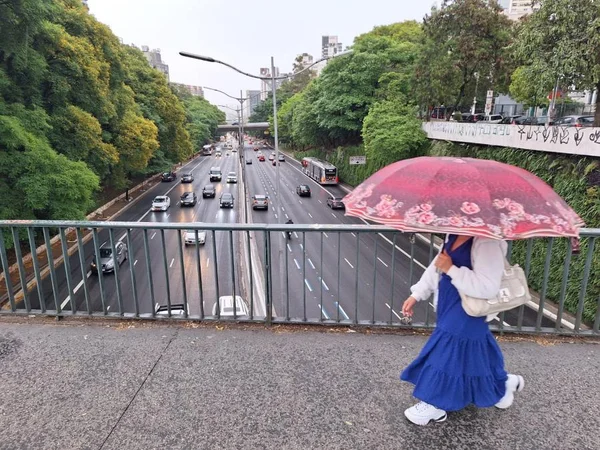 Imagem mostra mulher com saia azul, camisa clara e guarda-chuva rosa caminhando sobre viaduto - Metrópoles