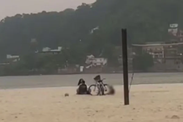 Imagem colorida de print de vídeo mostra praia ao fundo e, na areia, é possível ver uma bicicleta e duas pessoas sentadas, uma no colo e de frente para outra - Metrópoles