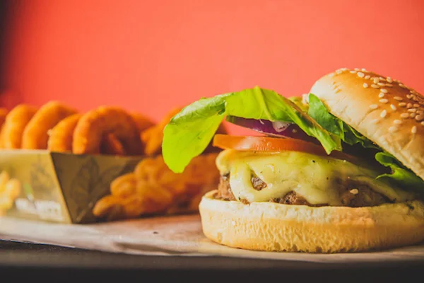 NA FOTO TEMOS UM HAMBURGUER COM CARNE, QUEIJO, CEBOLA, ALFACE E TOMATE E PAO E AO FUNDO ANEIS DE CEBOLA EMPANADOS