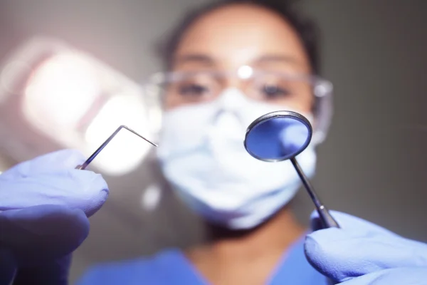 Imagem colorida mostra uma espátula e um espelho de dentistas, no primeiro plano, com uma dentista, mascarada, desfocada, ao fundo - Metrópoles