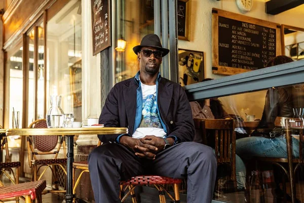 Foto colorida de Omar Sy, homem negro com casaco preto, chapéu e blusa branca - Metrópoles