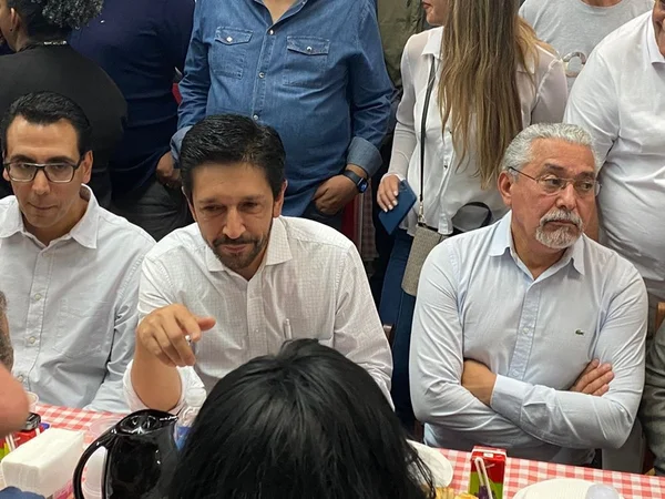 Imagem colorida mostra Ricardo Nunes, ao centro, sentado em uma mesa com pano xadrez e cercado de pessoas sentadas e em pé - Metrópoles