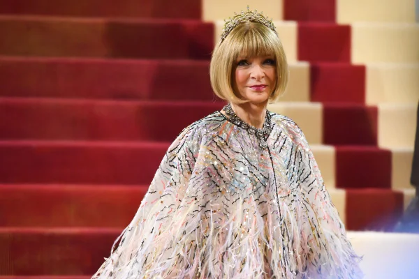 Anna Wintour, jornalista de moda e editora da revista Vogue, posando para foto no MET Gala de 2022. Ela é uma mulher idosa branca, com cabelo em corte chanel louro, e uma franja que cobre a testa. Veste uma roupa rosa brilhante, com detalhes de pluma e, na cabeça, uma tiara de diamantes.