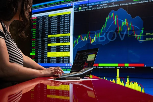 imagem colorida operadora da B3 com computador diante de gráfico do Ibovespa