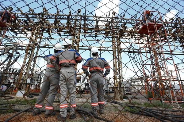 Fotografia colorida de funcionários uniformizados olhando para subestação de energia