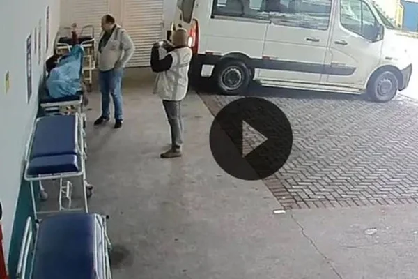 imagem colorida de dois homens tirando foto de um corpo em cima de uma maca, em um estacionamento