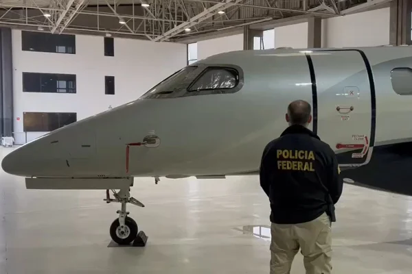 Foto colorida de homem de costas, virado para jatinho, vestido com jaqueta da Polícia Federal