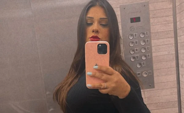 Mulher branca com cabelos escuros, batom vermelho e blusa preta segura celular em frente ao rosto. É empresária