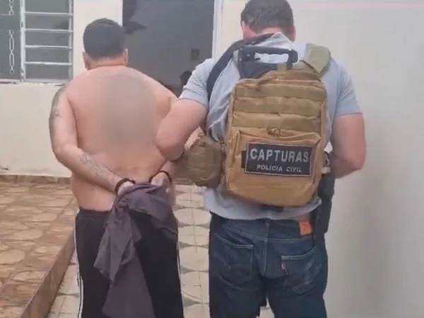 Em foto colorida homem acima do peso, sem camisa e de costas, do lado esquerdo, com as mãos algemadas para trás, sendo conduzido por policial civil usando colete balístico de cor clara - Metrópoles