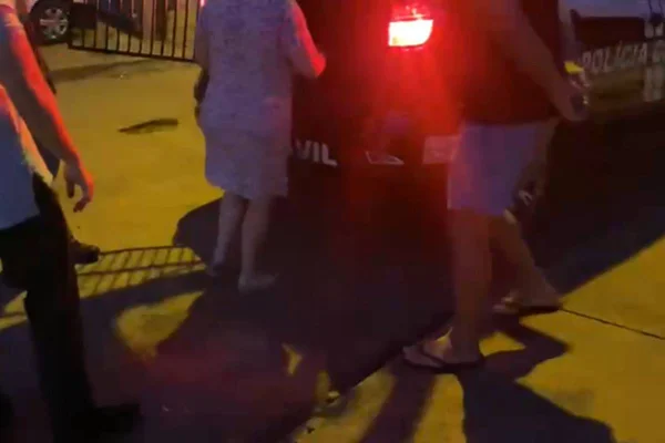 Foto colorida de casal de mulher e homem de pijamas sendo conduzidos, algemados, para carro de Polícia