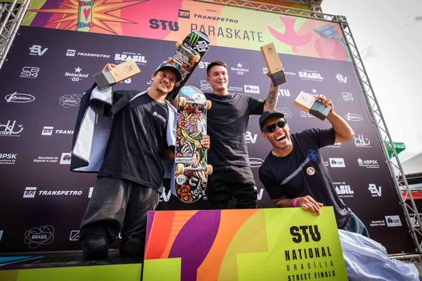 Três skatistas posam no pódio sorrindo, com skates erguidos e painel colorido ao fundo com logos do STU