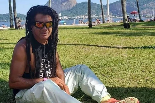 foto colorida de homem de dreads, oculos escuros, regata preta e calça branca sentado em gramado - metrópoles