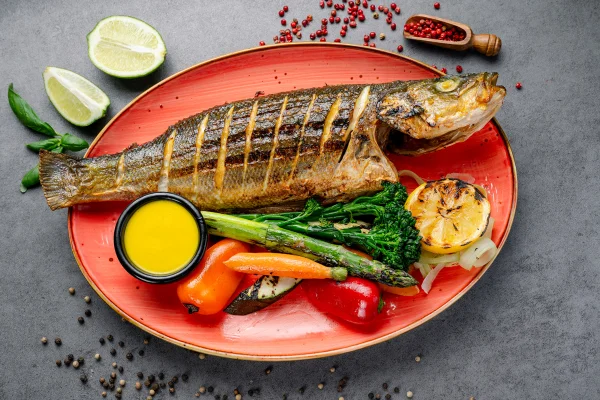 Foto colorida de peixe assado em prato com legumes - Metrópoles
