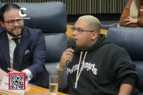 imagem colorida mostra mc ryan e seu advogado. ryan é negro, tem cabelo pintado de loiro, usa óculos e veste um moletom. ele segura um microfone. o advogado é branco, usa barba e tem cabelo preto.