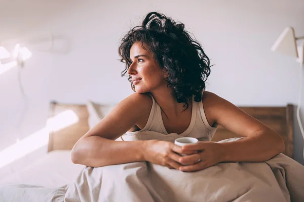 Foto colorida de mulher sentada em cama bebendo café - Metrópoles