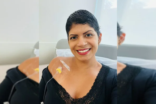 Mulher de cabelos negros e curtos posa para foto. Ela possui um eletrodo ligada à pele e está deitada em uma maca de hospital. Veste roupas pretas