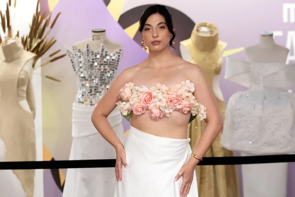 A imagem mostra uma mulher com um top de flores em frente roupas expostas em evento
