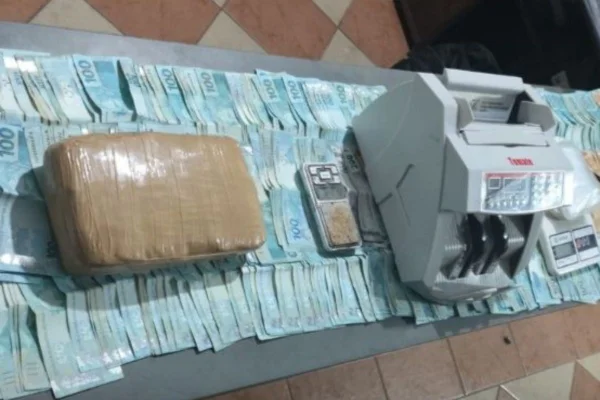 imagem colorida mostra notas de cem reais distribuídas em cima de uma mesa, junto com um pacote marrom e um equipamento para contar dinheiro