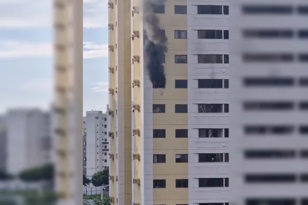 Imagem colorida de um prédio amarelo, com diversos apartamentos. Em um deles, está ocorrendo um incêndio, onde é possível ver uma fumaça preta saindo pela janela
